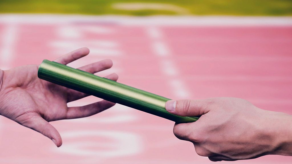 Primer plano de dos manos pasando un testigo de relevo en una pista de atletismo, ilustrando la transferencia de responsabilidad al negocio antes del go live.