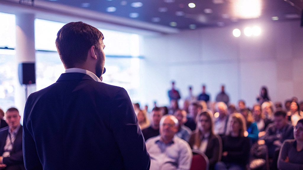 Orador corporativo de espaldas dirigiéndose a una audiencia numerosa en un auditorio luminoso, ilustrando una estrategia de comunicación interna para gestionar el cambio organizacional.