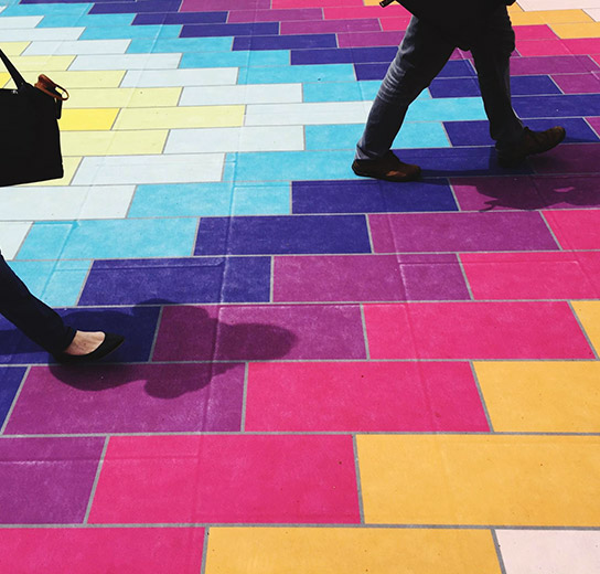 Personas caminando sobre un suelo pavimentado con bloques geométricos de colores brillantes