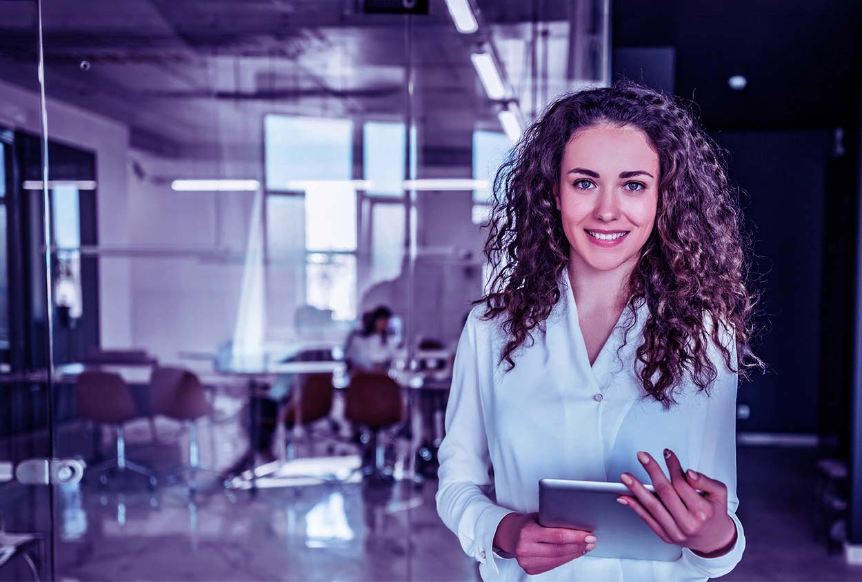 Una joven profesional sonriente con el cabello rizado, vestida con una blusa blanca y sosteniendo una tableta digital en una oficina moderna con salas de reuniones de vidrio al fondo