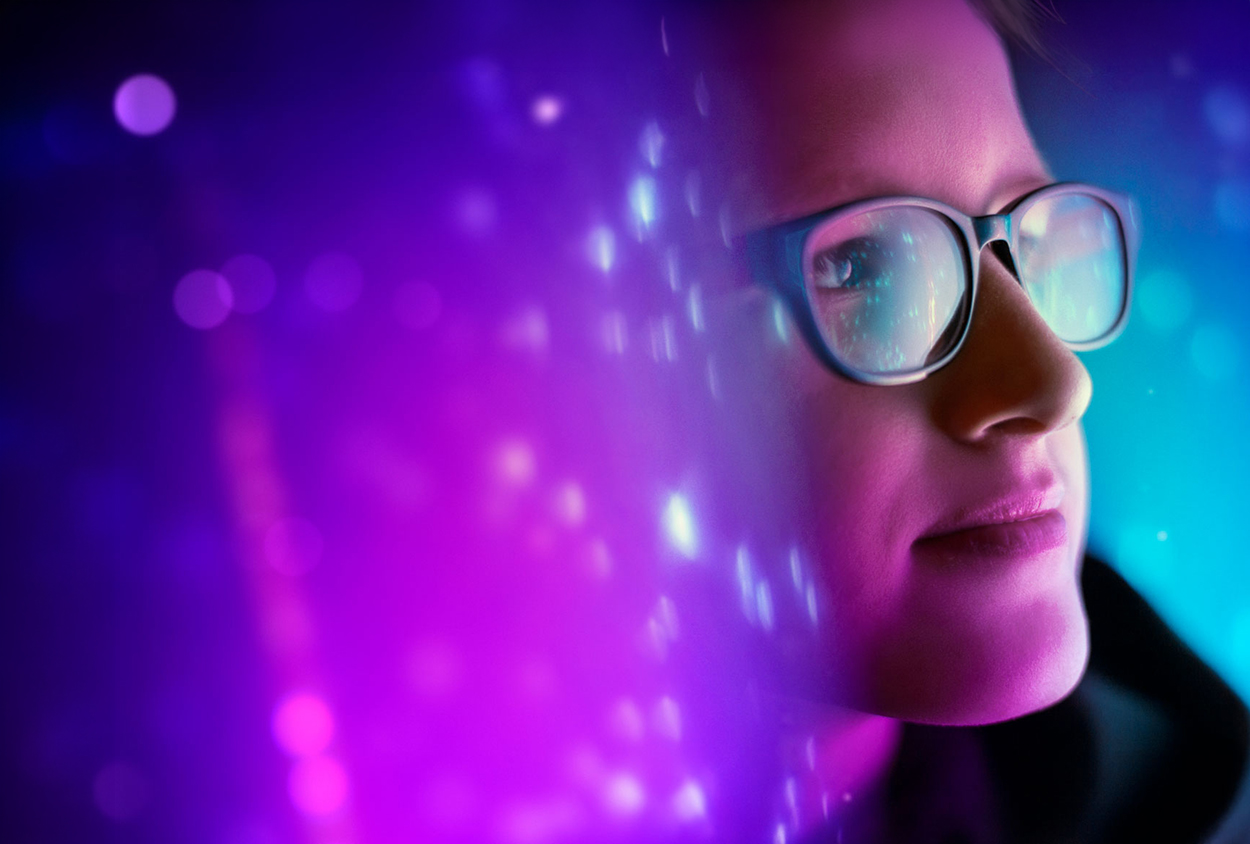 Primer plano de una persona con gafas mirando hacia el frente, iluminada por luces de neón en tonos morados y azules con efecto abstracto de desenfoque