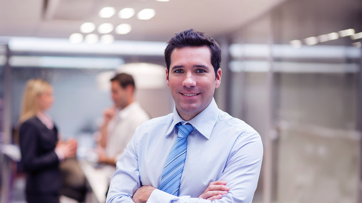 Primer plano de un hombre de negocios sonriendo con los brazos cruzados, vistiendo camisa clara y corbata azul a rayas. Al fondo, de manera difuminada, se observa una oficina moderna con dos colegas conversando