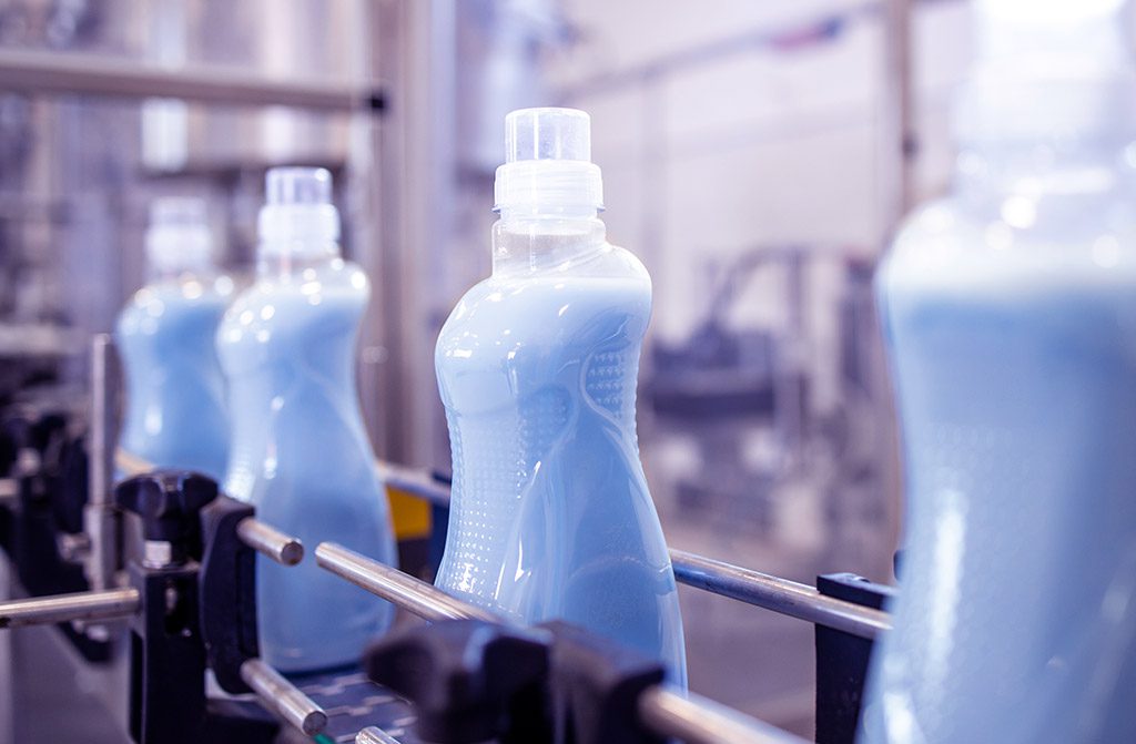 Botellas de detergente líquido azul avanzando en una cinta transportadora industrial automatizada.