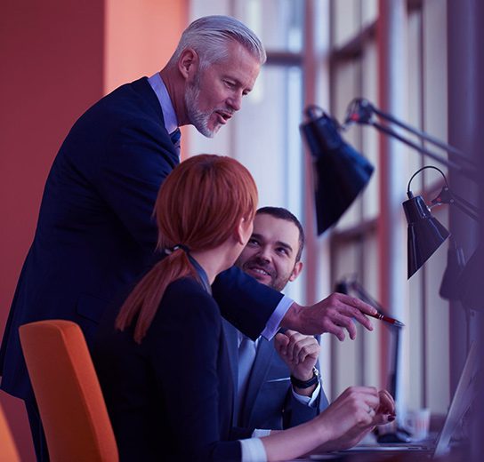 Ejecutivo senior capacitando y guiando a colaboradores frente a la computadora en un entorno de trabajo corporativo.